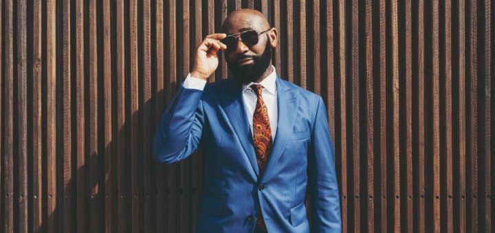 A man standing in front of a wooden wall wearing a blue suit with a white shirt and holding sunglasses to his face.