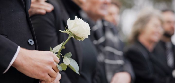 A close-up of a hand holding a white rose at a funeral in a cemetery. People in the background are in grief.