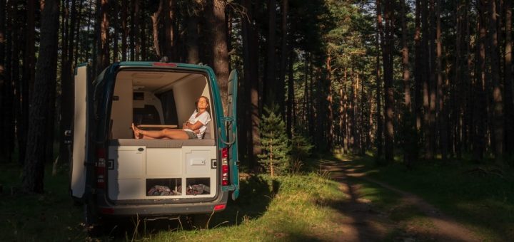 A young woman sits on a bed in her parked camper van and takes in the sun shining through the trees.