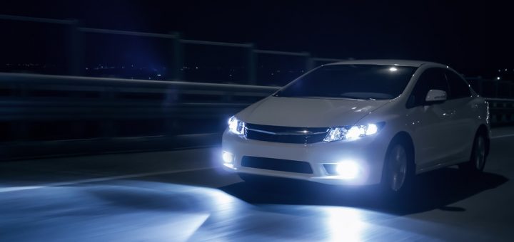 A white sedan driving down a road at night. The headlights are bright and illuminate the road in front of the car.