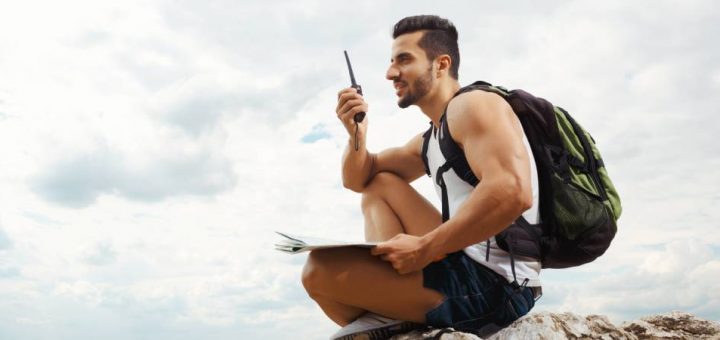 A man in a white tank top and denim shorts carrying a green backpack while sitting on a rock using a two-way radio.