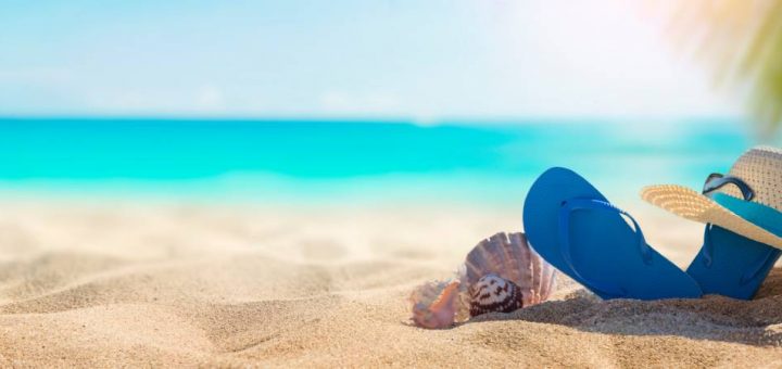 Blue flip-flops stick out of the sand, and a beach hat sits on one of the flip-flops. Shells sit next to the shoes.