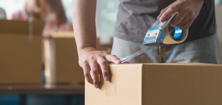 A man is taping up a cardboard box. There is a watch on his left wrist and a woman is out of focus in the background.