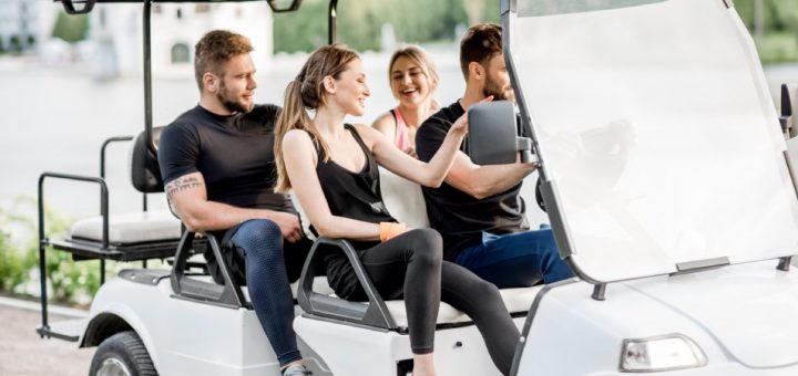 Four young adults in casual clothing drive down a paved road in a large white golf cart on a sunny day.