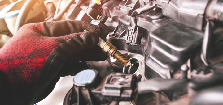An automotive mechanic holding a spark plug as they wear a red and black work glove. The plug is near the engine socket.