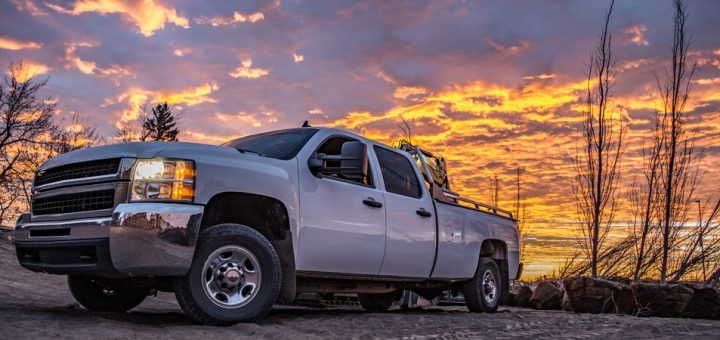 A heavy-duty diesel truck parked in front of a tree line. The truck is white and the sun is going down behind the trees.
