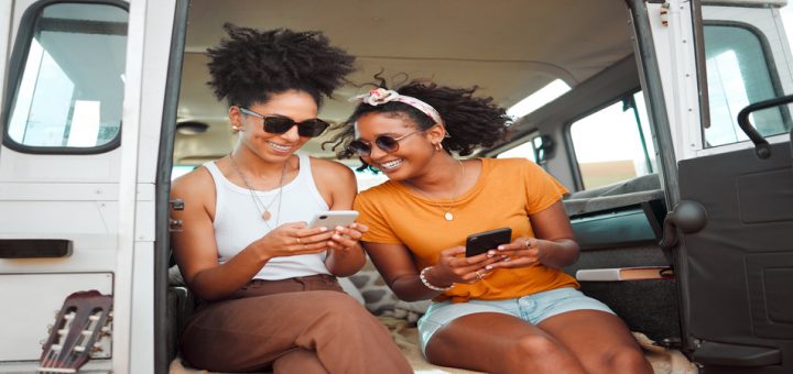 Two friends smiling and looking at each others phones while sitting on the floor of a white camper van.