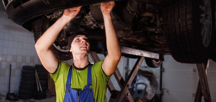 A man in blue overalls is working on the underside of a black car. The car is held up with a black scissor lift.