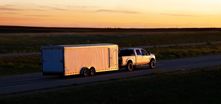 A white pickup truck pulls a white trailer down an empty rural highway. A beautiful sunset reflects off of the vehicles.