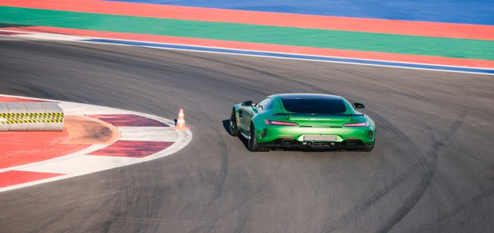 A green vehicle turns on a curved paved track near a traffic cone set on a white line. Tire marks are visible on the pavement.