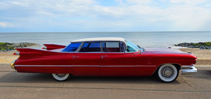 A vintage 1950s four door bright red Cadillac parked on the side of a road overlooking a beach and ocean.
