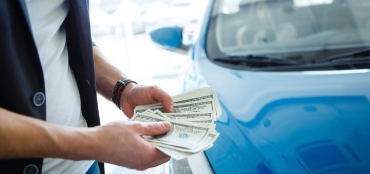 A man wearing a blue blazer stands next to a shiny blue car while fanning out a stack of money in both hands.