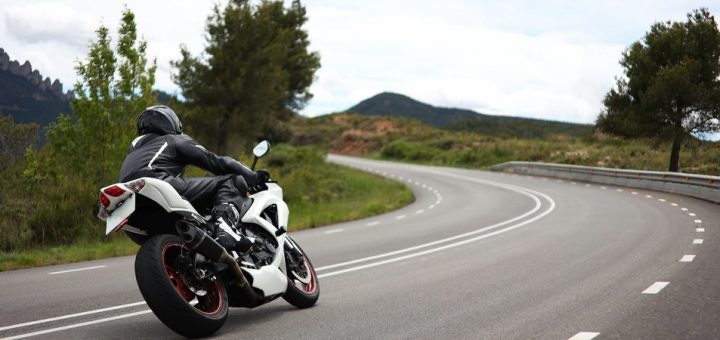 A person in leather attire and a motorcycle helmet riding a sleek white motorcycle and leaning in to turn a corner.