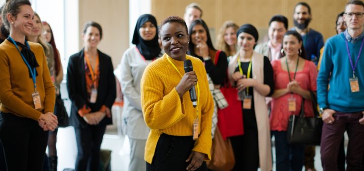 A woman wearing a yellow sweater using a wireless microphone with a group of people standing behind her.