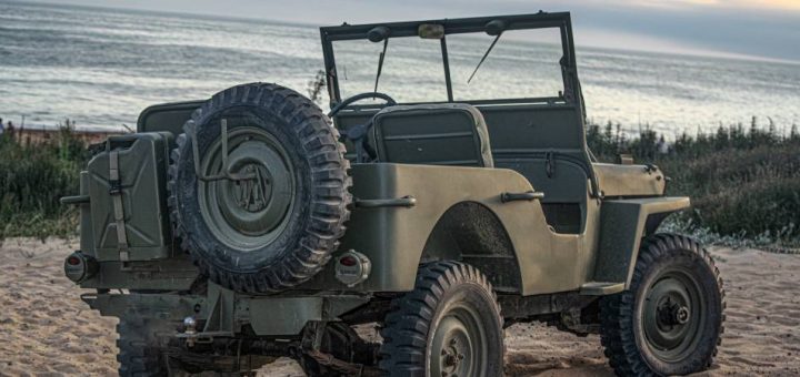 A dark green-colored vintage World War II era military Willys Jeep sits parked on the beach near an ocean.