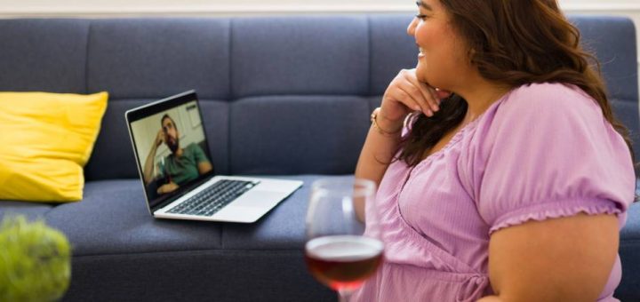 A woman in a purple shirt drinking a glass of red wine as she speaks to a man via a video call on her laptop.