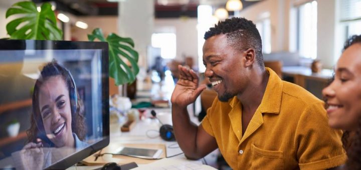 Two smiling people are sitting at a desk talking to a woman wearing a headset on a computer screen.