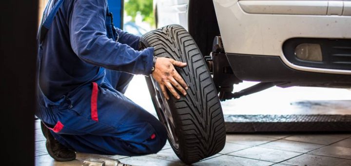 A mechanic is taking a tire off an elevated car. The mechanic is wearing a blue jumpsuit and has tools next to them.