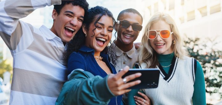 A diverse, fashionable friend group of younger adults is standing outside smiling and taking a selfie.