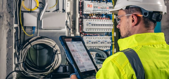 A man in a bright uniform, safety glasses, and a white hard hat looking at a tablet while standing in front of a switchboard.