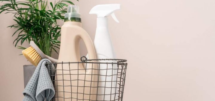 Plain cleaning product bottles in a basket with a cloth, brush, and a green plant, against a neutral background.