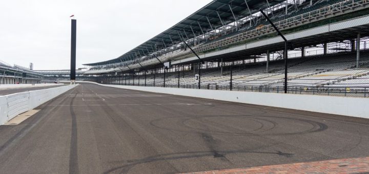 Tire tracks follow the track near the Indianapolis Yard of Bricks, with empty bleachers to the side and an overcast sky above.