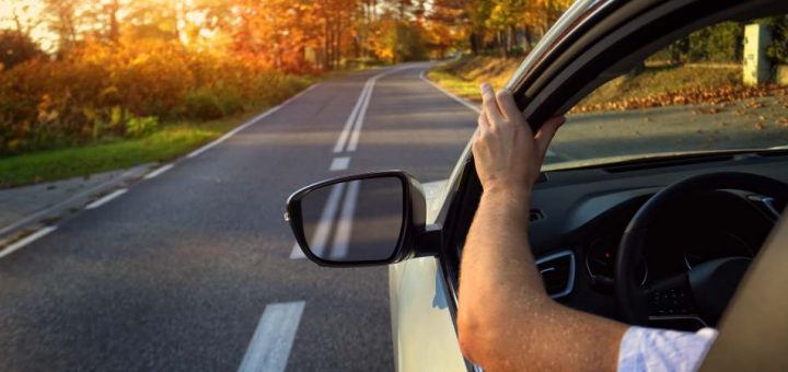 A motorist with their window down and arm placed on the open window. The road is curved with fall foliage all around.