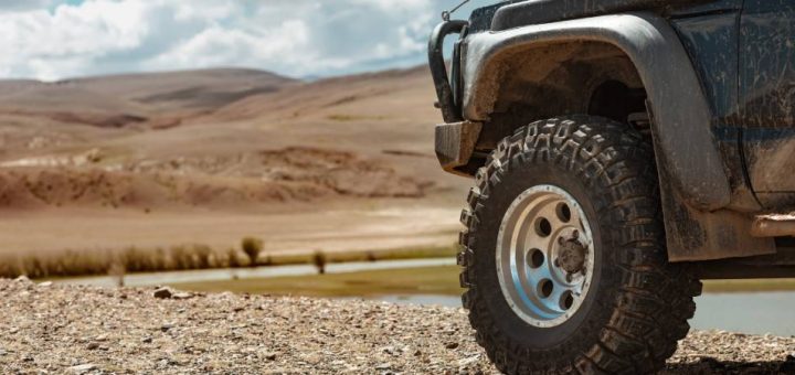 A close-up of the front left tire of an off-roading vehicle parked on a gravel surface, looking out over a dry landscape.