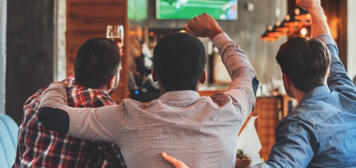 Three men huddled around a table raise their arms in celebration while watching a sports game on TV at a bar.