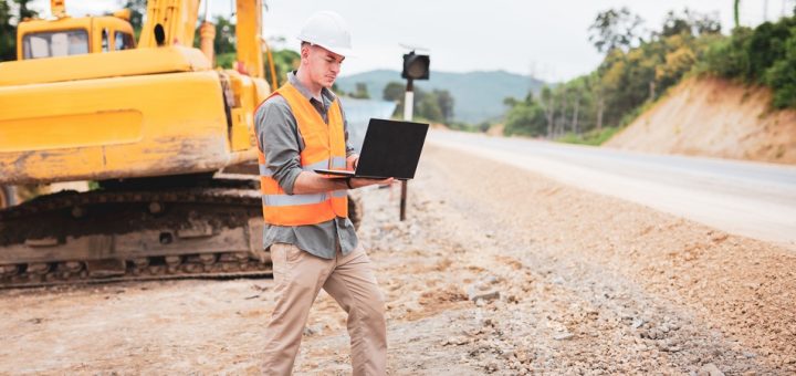 A mining operator using a laptop in front of a bulldozer. He is wearing an orange reflective vest and a white hard hat.