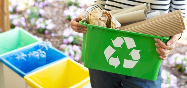 A close-up of a green basket with a recycling symbol, containing papers, held by a woman in a striped black and white top.
