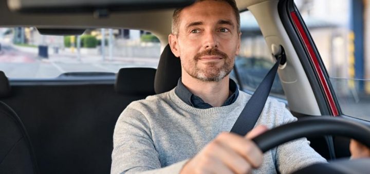 A mature man is slightly smiling as he looks in his rear-view mirror while driving his car on the road.