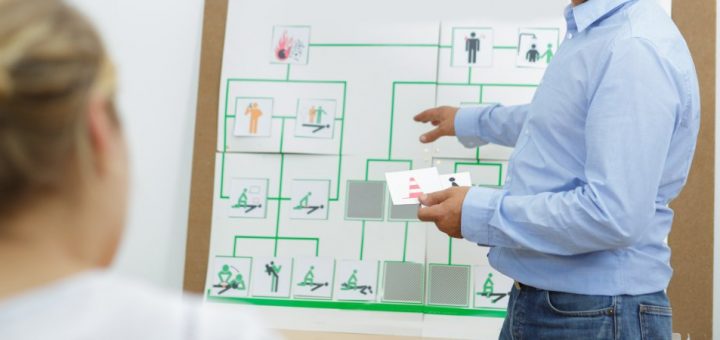 A man points to a display behind him with several emergency icons on it. Another person is blurred in the foreground.
