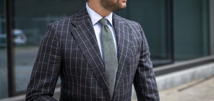 A bearded man wears a gray plaid suit jacket with a white button-down shirt and a wide gray tie outdoors.