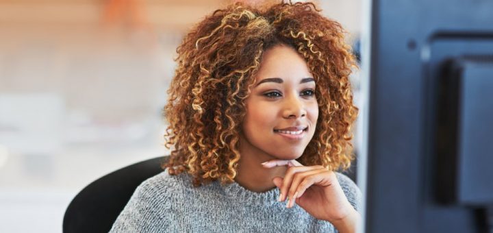 A smiling woman sits and looks at a computer monitor as she reads something. She wears a gray knit sweater.