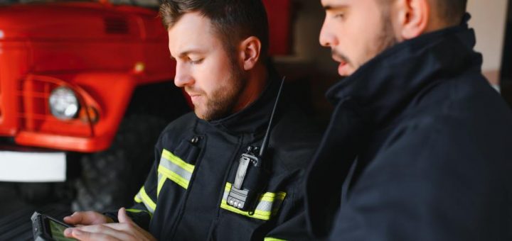 A pair of firefighters standing together beside a firetruck to review information on a handheld tablet.