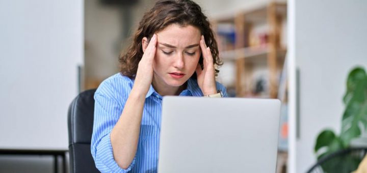 A young woman sits with her head in her hands, frustrated by poor computer speeds. She works from her home office.
