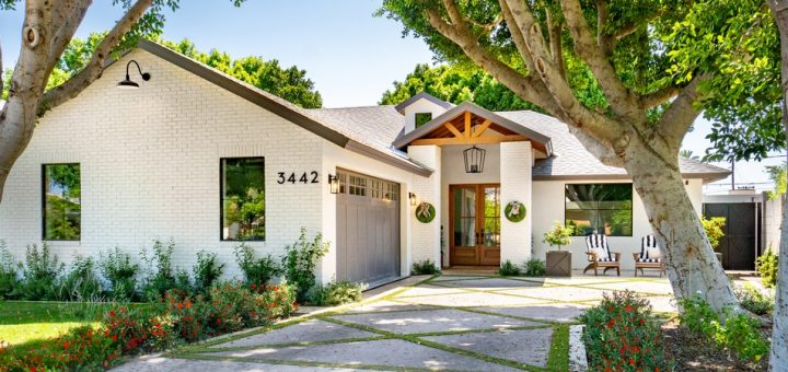 A modern white, single-story home with a grey garage door and brown front doors is situated behind a front yard with well-maintained flowers and trees.