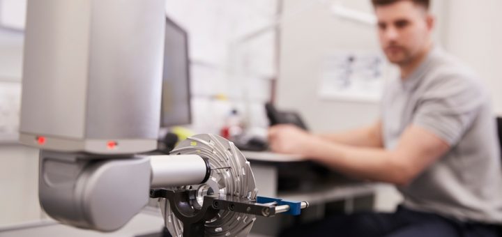 A coordinate measuring machine taking measurements of a small part. A man sits at a desk in the background.