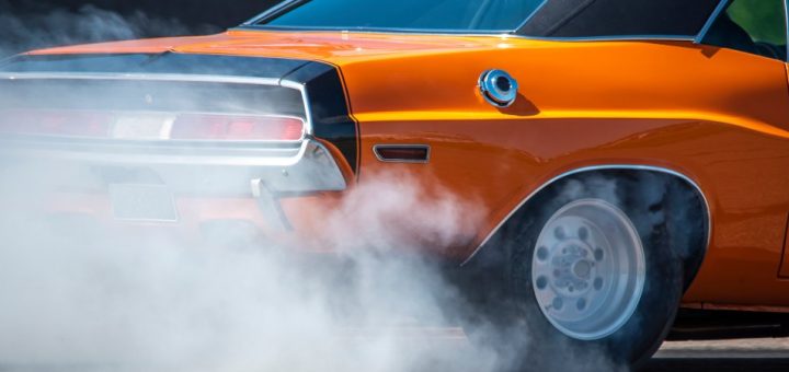The rear of an orange and black muscle car burns rubber, producing a plume of smoke from its back tires.