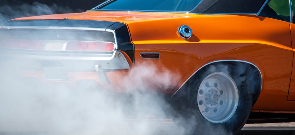 The rear of an orange and black muscle car burns rubber, producing a plume of smoke from its back tires.