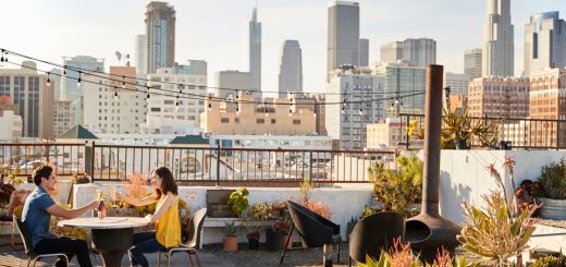 A man and woman toast with wine at a circular rooftop table, with the city skyline in the background.