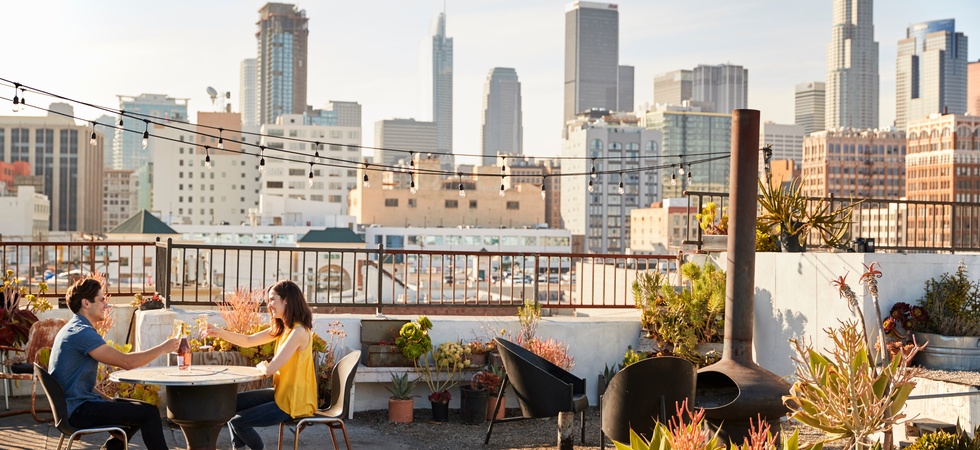 A man and woman toast with wine at a circular rooftop table, with the city skyline in the background.