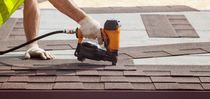 A person in a yellow safety vest uses a black and orange nail gun to install brown shingles on a house.