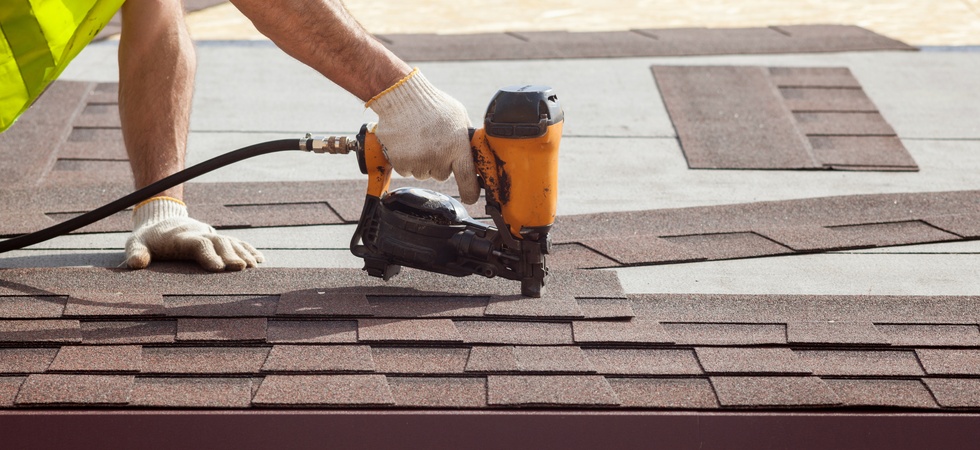 A person in a yellow safety vest uses a black and orange nail gun to install brown shingles on a house.