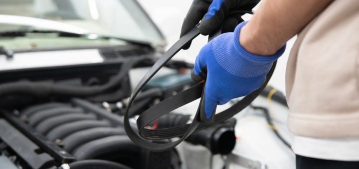A man wearing blue safety gloves stands in front of the open hood of a vehicle and its engine, holding an engine belt.
