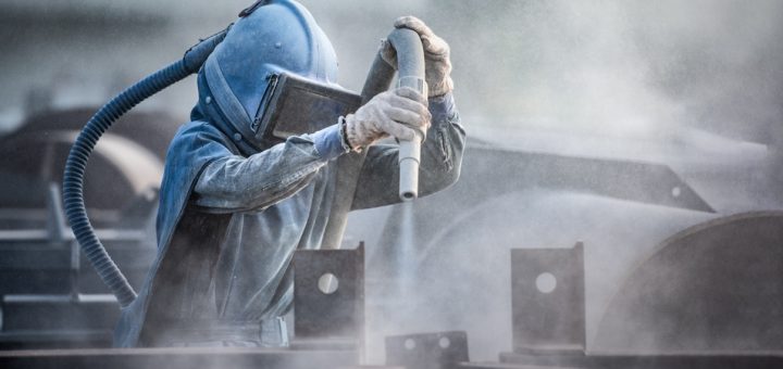 A worker in a comprehensive personal protective suit uses a sand blaster to prepare steel parts at a factory.