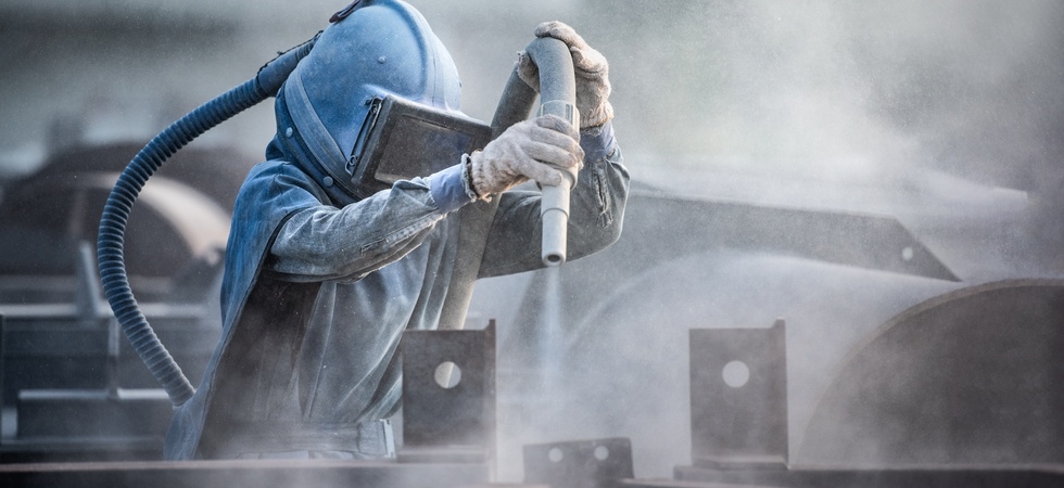 A worker in a comprehensive personal protective suit uses a sand blaster to prepare steel parts at a factory.