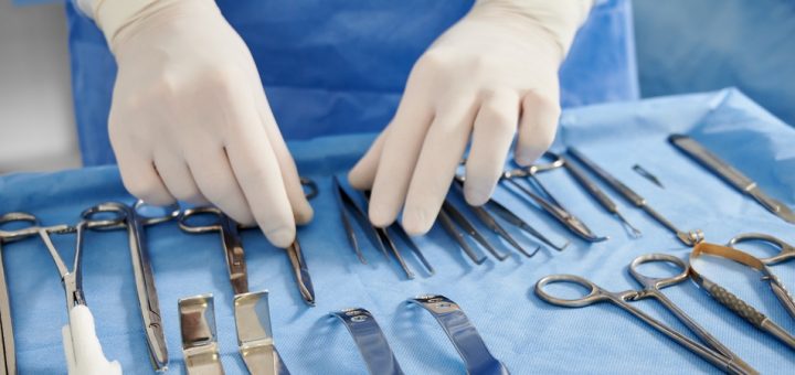 An individual in blue scrubs and white gloves organizing stainless steel surgical instruments on a tray.