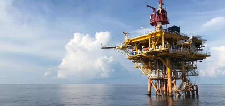 An oil rig in the middle of the water, there is no land in sight around it. The sky is blue with some cloud coverage.